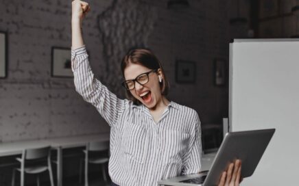 CREATIVITA MEDITAZIONE MINDFULLNESS Donna in camicia a righe che festeggia con il pugno alzato mentre utilizza un laptop.